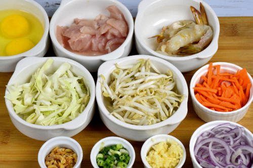 List of ingredients to cook the Singapore noodles. From top left to right bottom: eggs, chicken, shrimps, cabbage, bean sprouts, carrots, dry shrimps, scallions, garlic, onions.